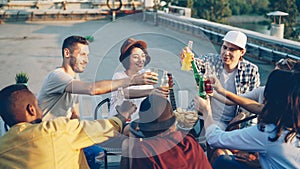 Cheerful friends are chatting and drinking lemonade sitting on rooftop on warm sunny day with snacks and drinks on table