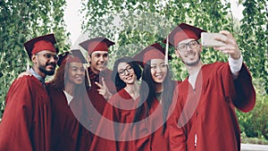 Cheerful friends graduating students are taking selfie with smartphone, young people are posing, showing hand gestures