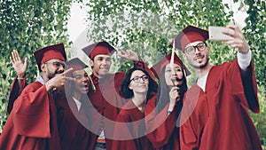 Cheerful friends graduating students are taking selfie with smartphone, young people are posing, showing hand gestures