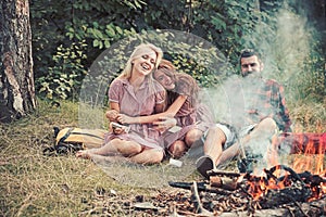 Cheerful friends having picnic in woods. Bearded man and two smiling girls reading books by campfire, sustainable