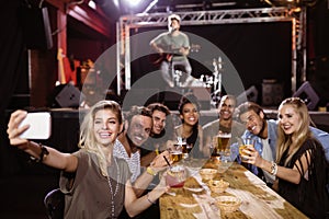 cheerful friends taking selfie while sitting at table with performer singing on stage