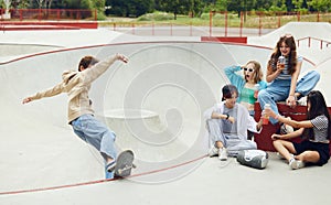 Cheerful friends, teens meeting together at skateboard park, having fun, riding on skate, talking. Boy doing stunts