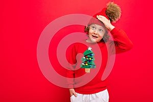Cheerful funny boy on a red background in a warm hat and sweater with a Christmas tree