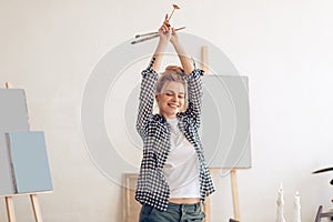 Cheerful funny girl with hairbun raising her arms and holding brushes