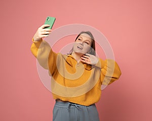 Cheerful funny woman 40s wearing casual sweater posing doing selfie shot on mobile phone over pink background