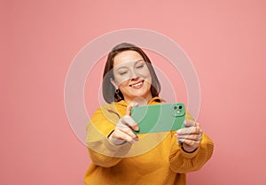 Cheerful funny woman 40s wearing casual sweater posing doing selfie shot on mobile phone over pink background