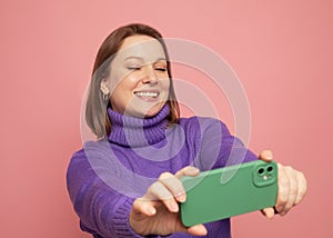 Cheerful funny woman 40s wearing casual sweater posing doing selfie shot on mobile phone over pink background