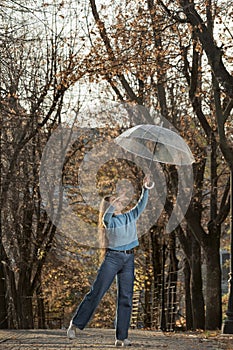 Cheerful girl in blue sweater and pants with transparent umbrella on alley. Beautiful woman dance in autumn park
