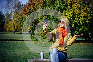 Cheerful girl in bright clothes and sunglasses sitting on park bench and taking selfie on smartphone against autumn trees.