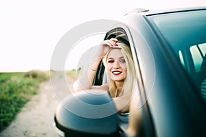 Cheerful girl is driving her new car with pleasure. The lady raises her elbow through the window and touching her hair gently