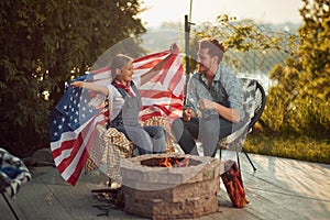 Cheerful girl and father sitting outdoors by a fireplace in their yard roasting marshmallows, holding USA flag