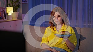 cheerful girl in glasses laughing from watching funny program on TV, eating fast food at night while sitting on couch in