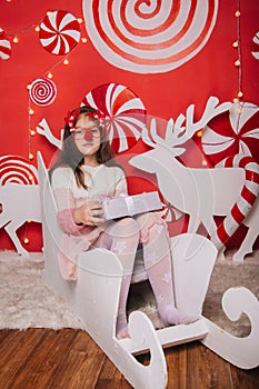 A cheerful girl in glasses with a red nose and horns is holding a gift in her hands. A wall of lollipops. The child is sitting in