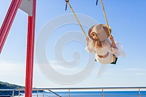 Cheerful girl in a hat and dress riding on a swing overlooking the sea.