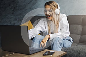 Cheerful girl in headphones sitting on sofa in front of laptop. Entertainment online content for streaming at affordable