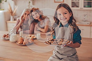 Cheerful girl holding plate with biscuits and looking at the camera with mother and grandmother sitting at the kitchen table on