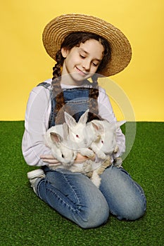 Cheerful girl in a jeans and straw hat holding a real three white rabbits,  yellow background. Vertical view.