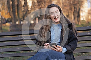 Cheerful girl with long brown hair sits in park on bench and uses her phone. Young woman with smartphone outside