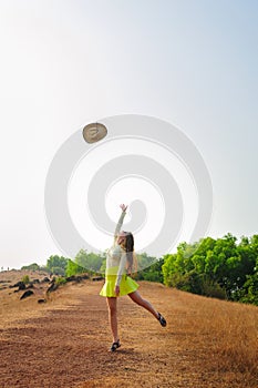 Cheerful girl with long hair jumps and throws straw hat in the air. Young brunette woman in short skirt walks on hill