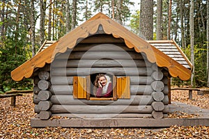 The cheerful girl looks out the window of a wooden log house and talks on a smartphone
