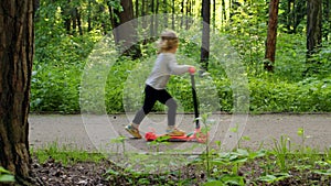 cheerful girl on orange scooter drives past camera in park in sunny and summer forest. small teenage child kid drive