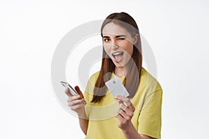 Cheerful girl pays on phone, shopping online with smartphone, showing credit card and winking, got cashback for purchase