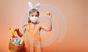 A cheerful girl with rabbit ears on her head and a protective mask with a basket of colored eggs in her hands
