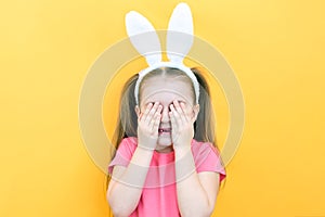 Cheerful girl with rabbit ears on her head on a yellow background. Funny crazy happy child. Easter child. Covers his eyes with his