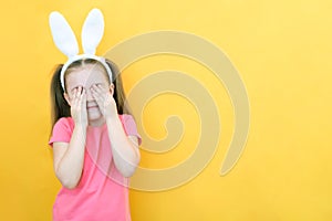 Cheerful girl with rabbit ears on her head on a yellow background. Funny crazy happy child. Easter child. Covers his eyes with his