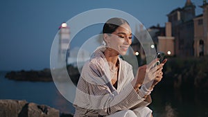 Cheerful girl reading message on smartphone at evening shore. Woman looking cell