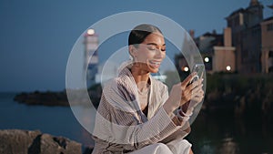 Cheerful girl reading message on smartphone at evening shore. Woman looking cell