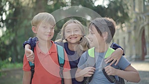 Cheerful girl running along sunny schoolyard and hugging boys. Portrait of three joyful Caucasian children with