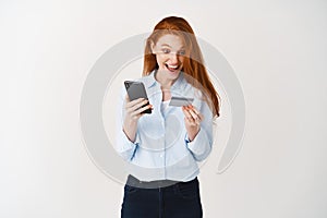 Cheerful girl shopping online, making purchase in smartphone app with plastic credit card, standing against white