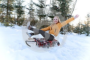 Cheerful girl sits on wooden sled on pines or fir trees background. Active weekend Outside in the winter forest