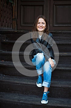 A cheerful girl in sneakers, jeans and a leather jacket sits on