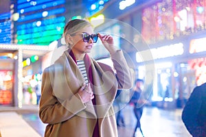 Cheerful girl in trendy apparel excited with night lights in Big City, beautiful brunette woman in spectacles happy