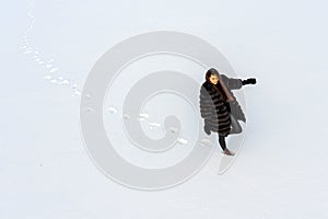 A cheerful girl in a warm fur coat walks across the open field leaving footpath in the snow