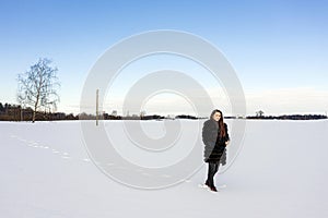 A cheerful girl in a warm fur coat walks across the open field leaving footpath in the snow