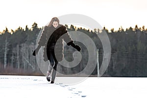 A cheerful girl in a warm fur coat walks across the open field leaving footpath in the snow