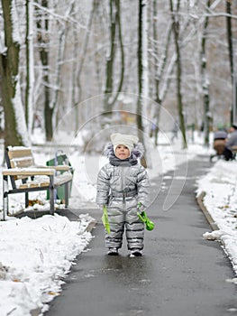 A cheerful girl in warm silver overalls is standing on the path in winter