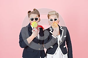 Cheerful girls couple with lollipops and glasses. Girls hide mouth by lollipops and eyes by black sun glasses.