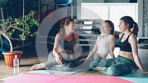 Cheerful girls are socialising in wellness center sitting on yoga mats on wooden floor. Sports equipment, green plant