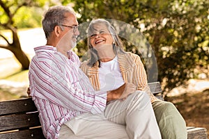 Cheerful glad european mature couple sit on bench, enjoy date together in park, talk at weekend in summer