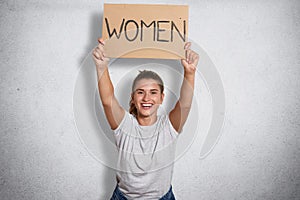 Cheerful good looking feminist smiling sincerely, raising inscription women in both hands, ready to public activity, peaceful