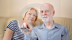 Cheerful good-looking senior couple sitting on sofa at home. Watching television, switching channels