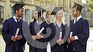 Cheerful graduate students in academic regalia talking to each other, success