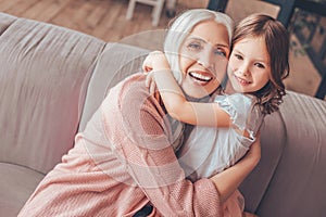 Cheerful grandmother hugging granddaughter sitting on the sofa in living room and looking at the camera