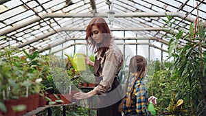 Cheerful greenhouse worker is watering plants in workplace with her helpful daughter and talking to child while little