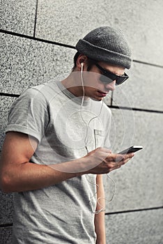 Cheerful guy dressed in gray t-shirt, sunglasses and hat at the street, listening to music with earphones, holding mobile phone.