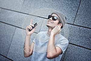 Cheerful guy dressed in gray t-shirt, sunglasses and hat at the street, listening to music with earphones, holding mobile phone.